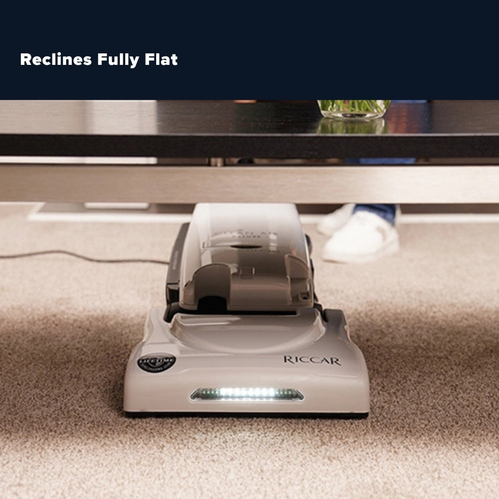 A Riccar under-desk mat shown on a carpeted floor under a table, with a dark overlay at the top reading 'Reclines Fully Flat'.